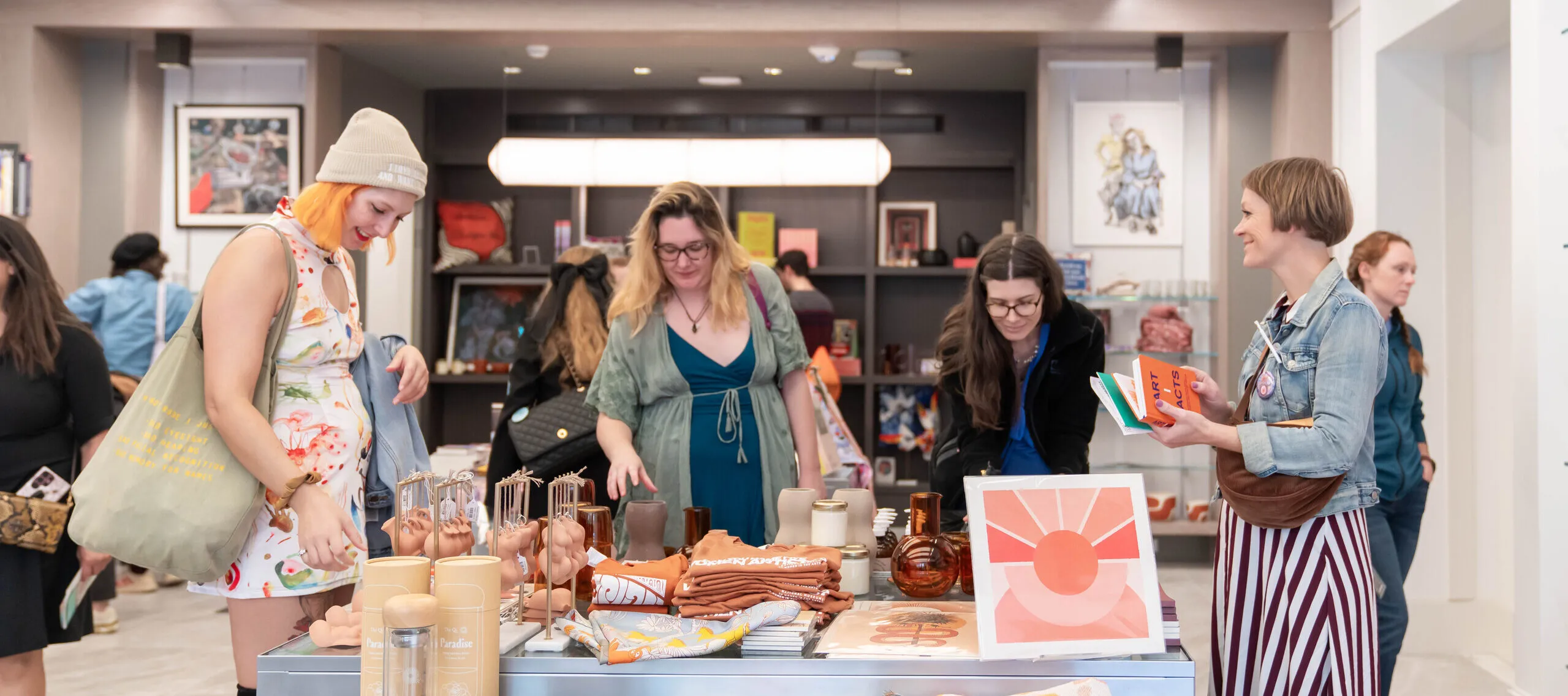 A group of women are standing in a shop around a table with colorful books, prints, and other objects on it. They have light skin tones and are wearing colorful dresses.