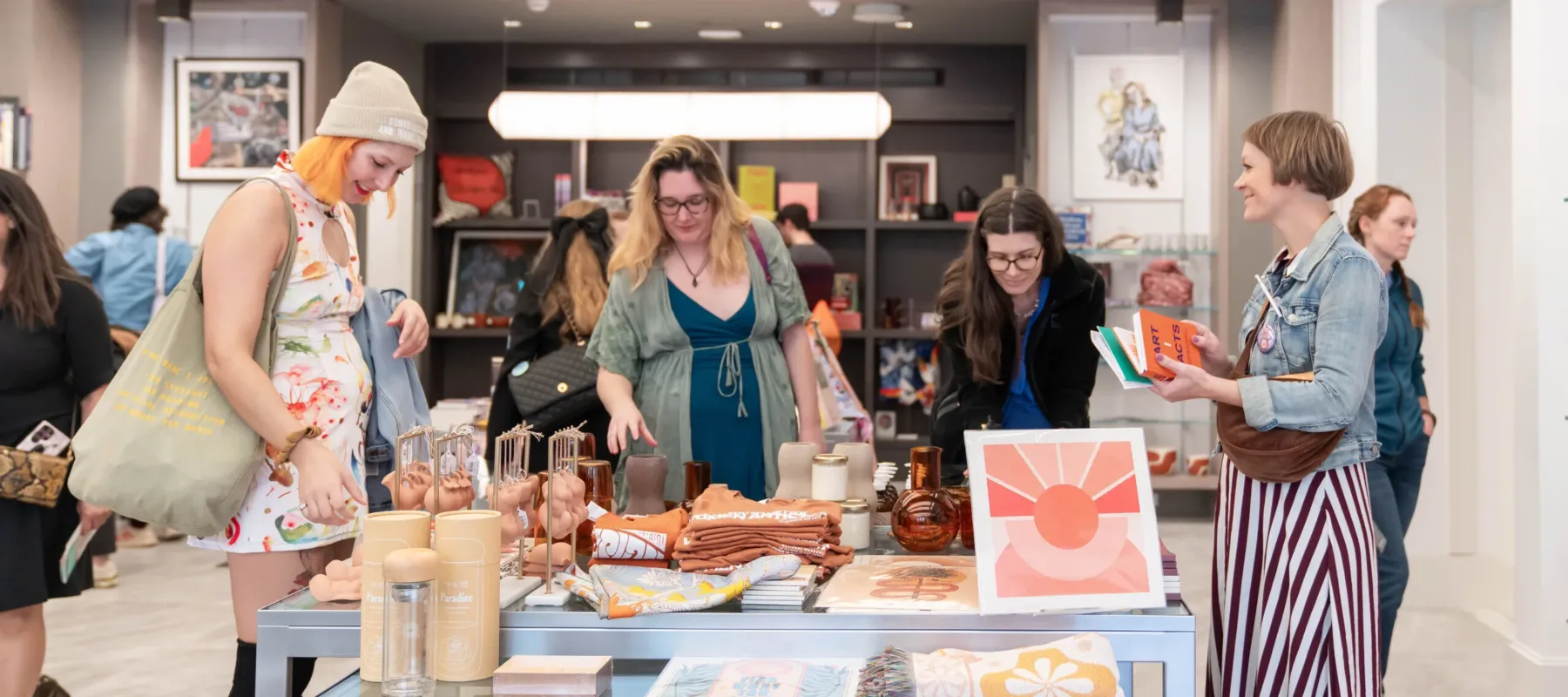 A group of women are standing in a shop around a table with colorful books, prints, and other objects on it. They have light skin tones and are wearing colorful dresses.