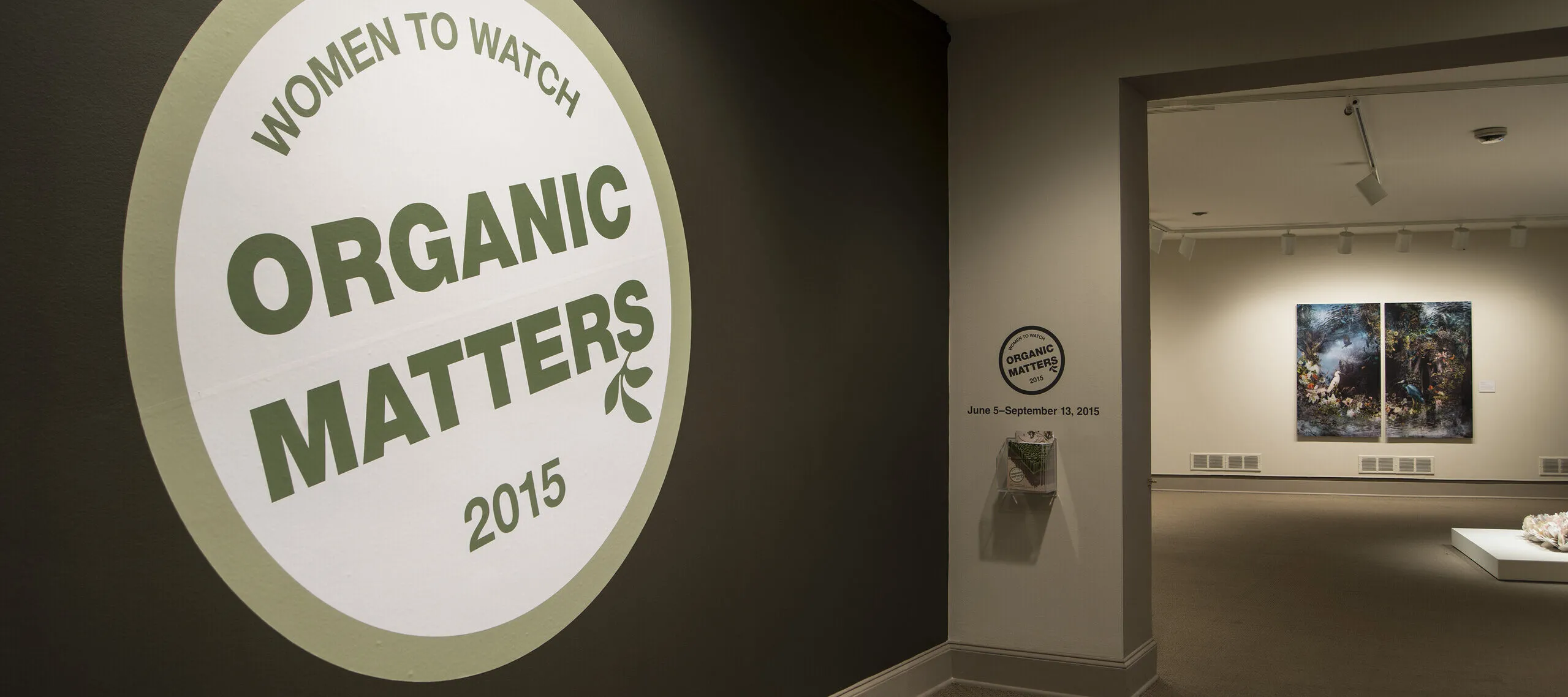 View of a gallery space. In big letters green , the text on the wall reads "Women to Watch: Organic Matters, 2015."