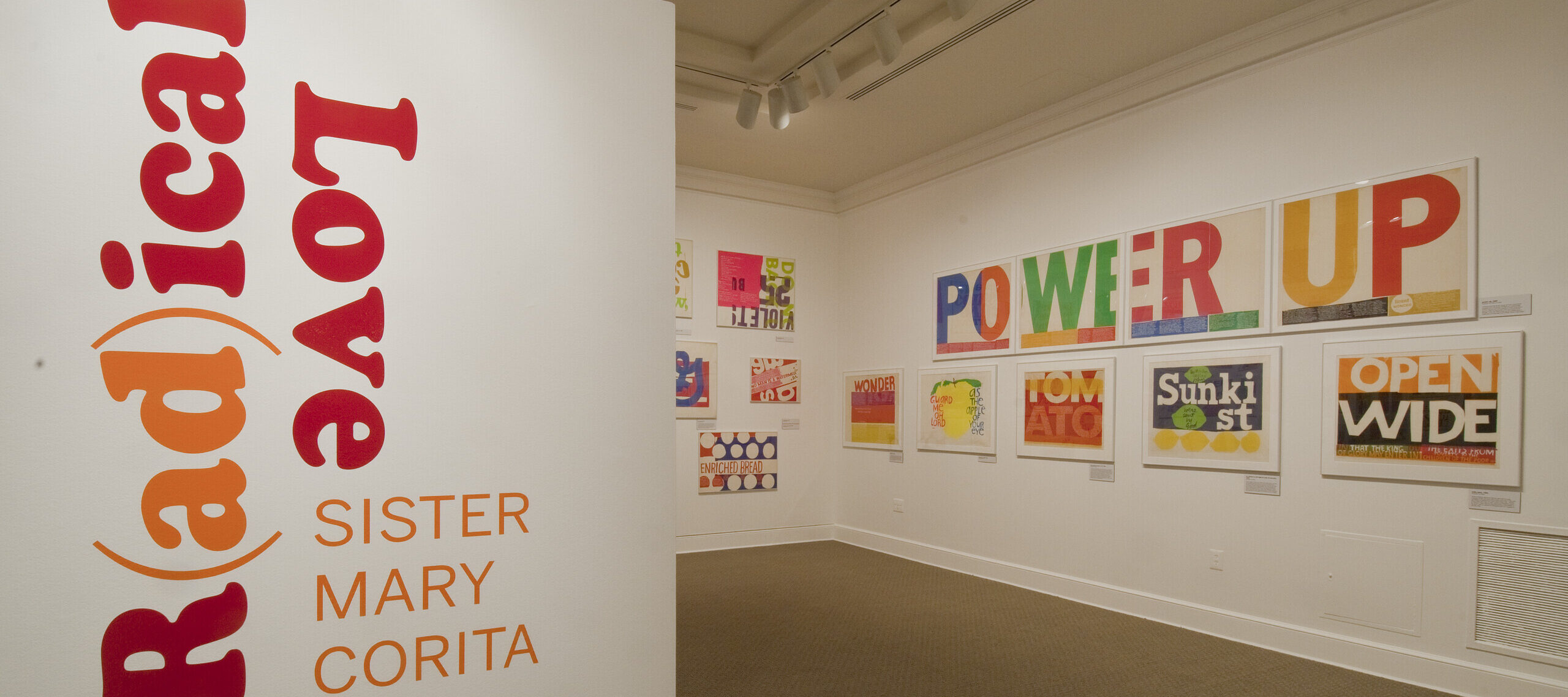 A white room with a wall in the foreground that has bold, red and orange text that reads ‘R(ad)ical Love: Sister Mary Corita, March 9–July 15, 2012.’ In the background, bold and colorful prints with text are hang on a white wall.
