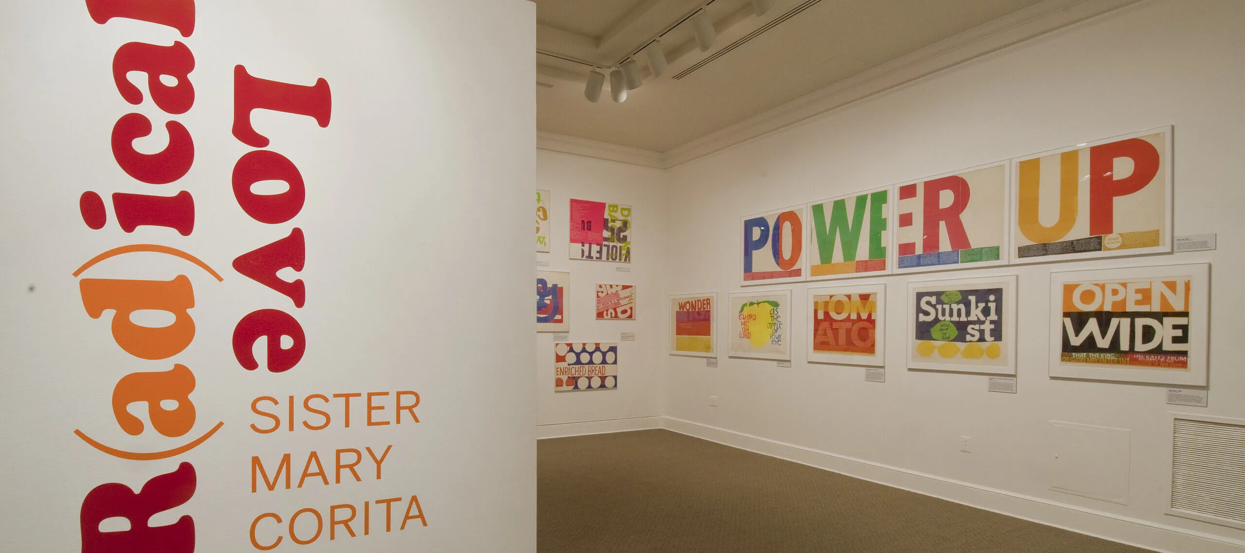 A white room with a wall in the foreground that has bold, red and orange text that reads ‘R(ad)ical Love: Sister Mary Corita, March 9–July 15, 2012.’ In the background, bold and colorful prints with text are hang on a white wall.