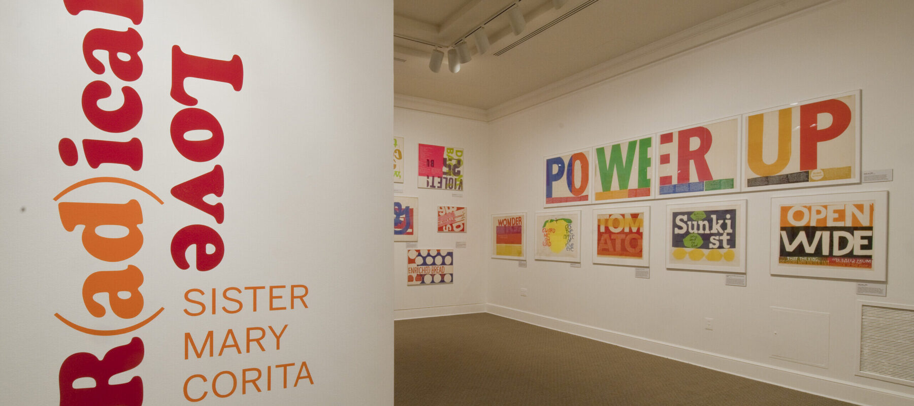 A white room with a wall in the foreground that has bold, red and orange text that reads ‘R(ad)ical Love: Sister Mary Corita, March 9–July 15, 2012.’ In the background, bold and colorful prints with text are hang on a white wall.