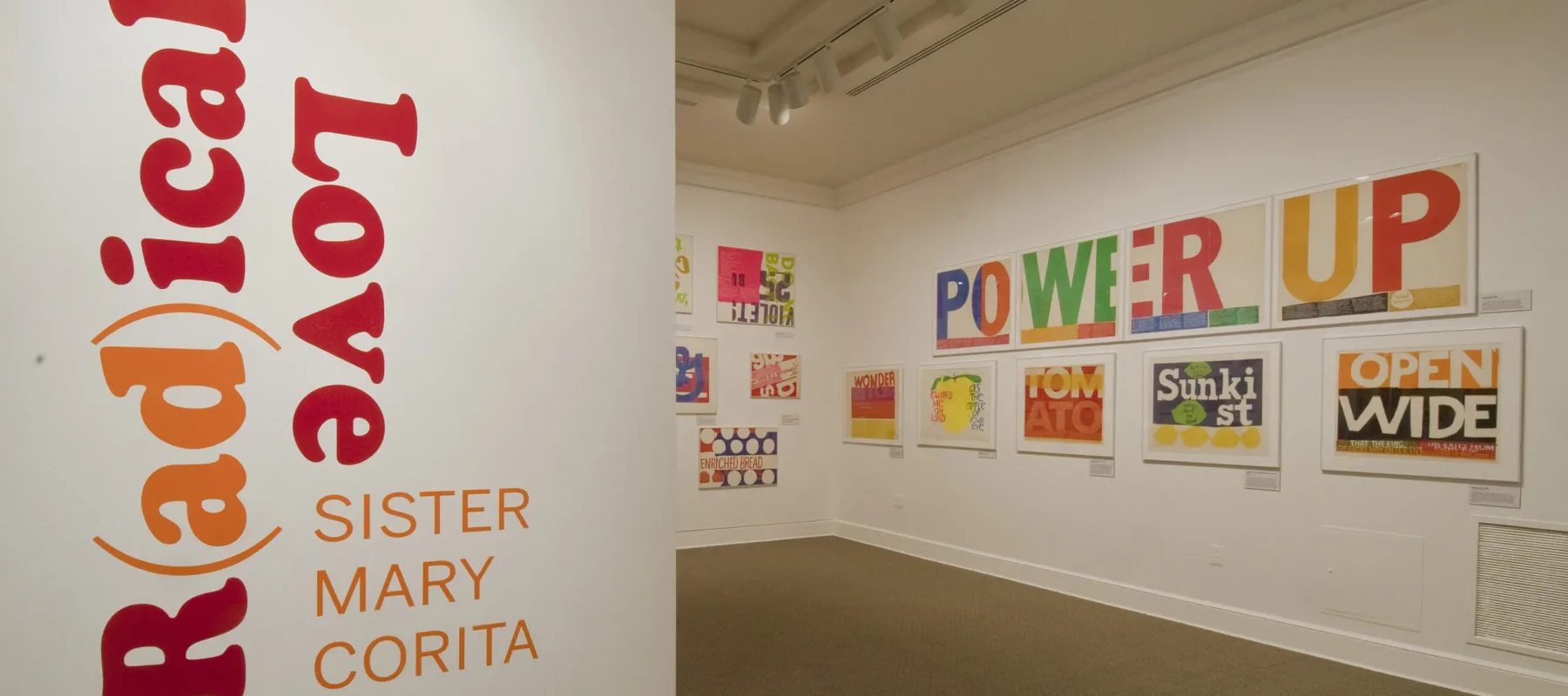 A white room with a wall in the foreground that has bold, red and orange text that reads ‘R(ad)ical Love: Sister Mary Corita, March 9–July 15, 2012.’ In the background, bold and colorful prints with text are hang on a white wall.