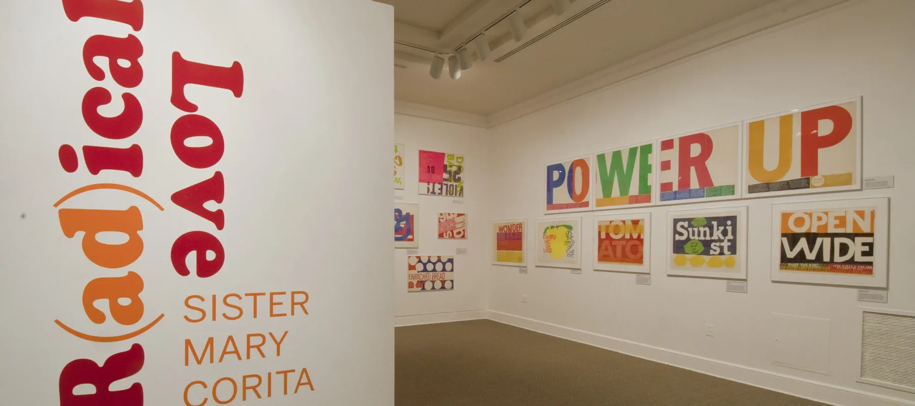 A white room with a wall in the foreground that has bold, red and orange text that reads ‘R(ad)ical Love: Sister Mary Corita, March 9–July 15, 2012.’ In the background, bold and colorful prints with text are hang on a white wall.