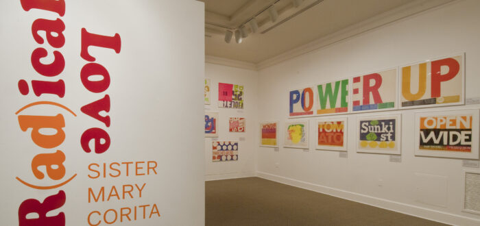 A white room with a wall in the foreground that has bold, red and orange text that reads ‘R(ad)ical Love: Sister Mary Corita, March 9–July 15, 2012.’ In the background, bold and colorful prints with text are hang on a white wall.