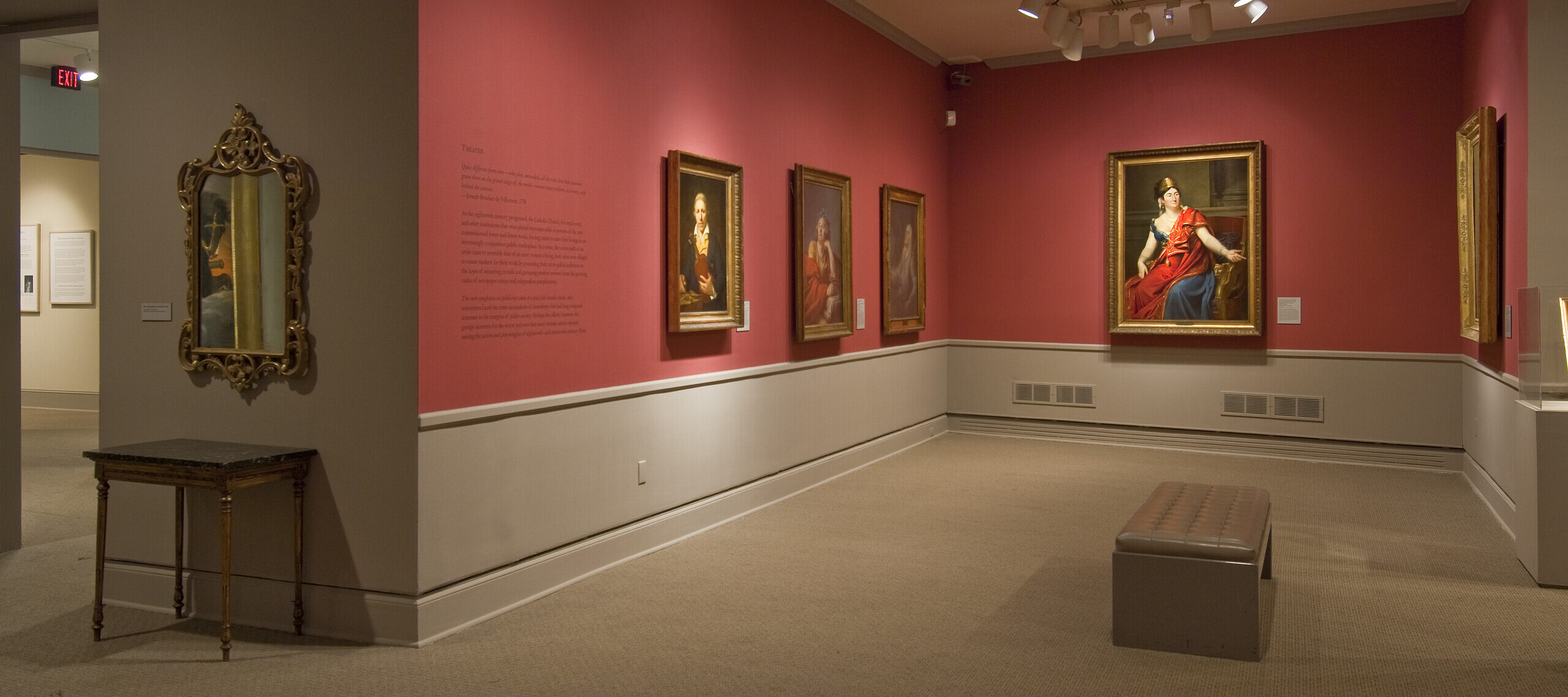 View of a gallery space with red walls. A golden mirror with plenty of ornaments is hanging to the left, large historical portraits of women are hanging to the right.