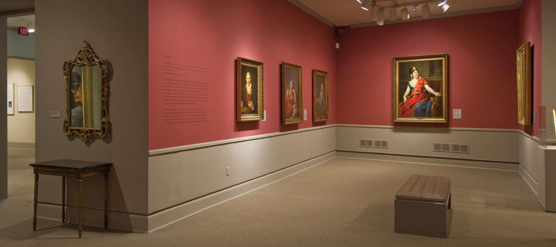 View of a gallery space with red walls. A golden mirror with plenty of ornaments is hanging to the left, large historical portraits of women are hanging to the right.
