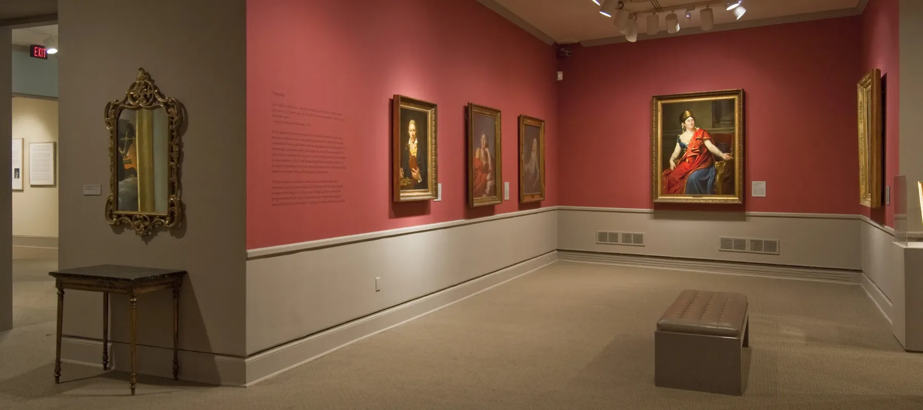 View of a gallery space with red walls. A golden mirror with plenty of ornaments is hanging to the left, large historical portraits of women are hanging to the right.