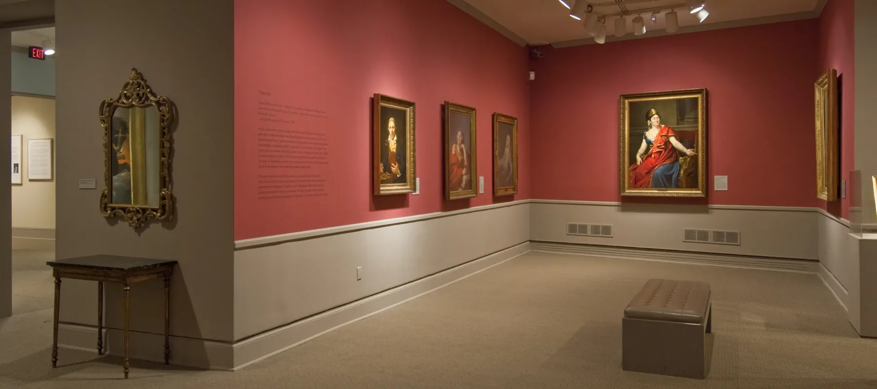 View of a gallery space with red walls. A golden mirror with plenty of ornaments is hanging to the left, large historical portraits of women are hanging to the right.