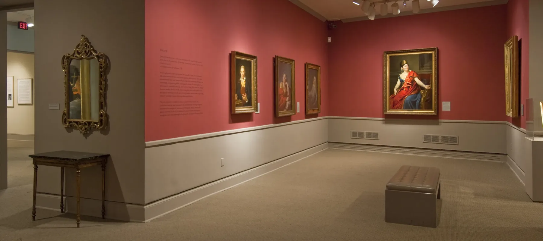 View of a gallery space with red walls. A golden mirror with plenty of ornaments is hanging to the left, large historical portraits of women are hanging to the right.