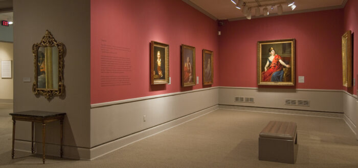 View of a gallery space with red walls. A golden mirror with plenty of ornaments is hanging to the left, large historical portraits of women are hanging to the right.