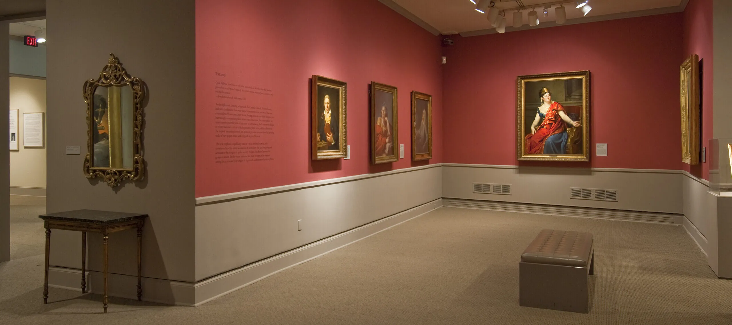 View of a gallery space with red walls. A golden mirror with plenty of ornaments is hanging to the left, large historical portraits of women are hanging to the right.