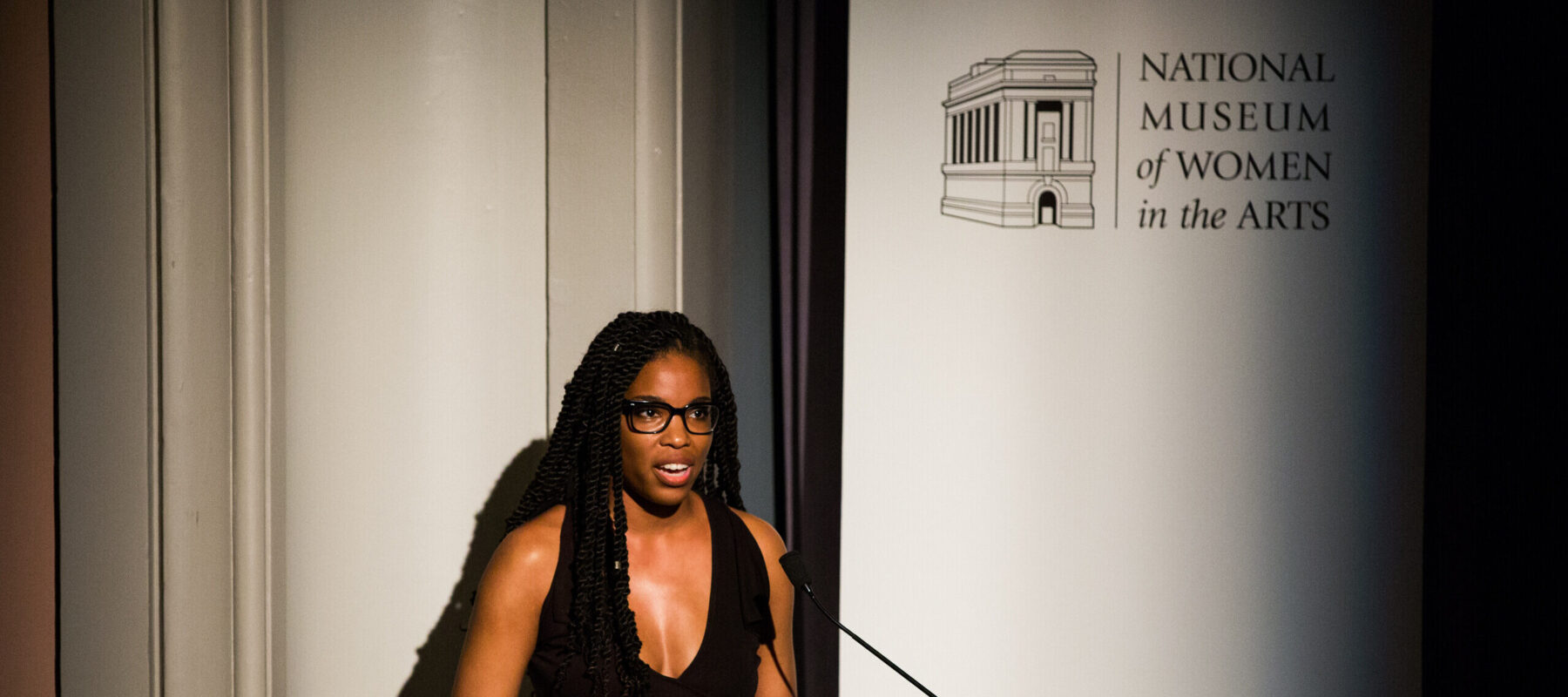 A woman with a dark skin tone is standing in front of a white background giving a speech by a wooden podium. On a banner, the text reads "National Museum of Women in the Arts."