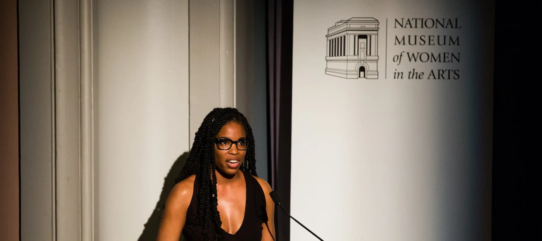 A woman with a dark skin tone is standing in front of a white background giving a speech by a wooden podium. On a banner, the text reads "National Museum of Women in the Arts."