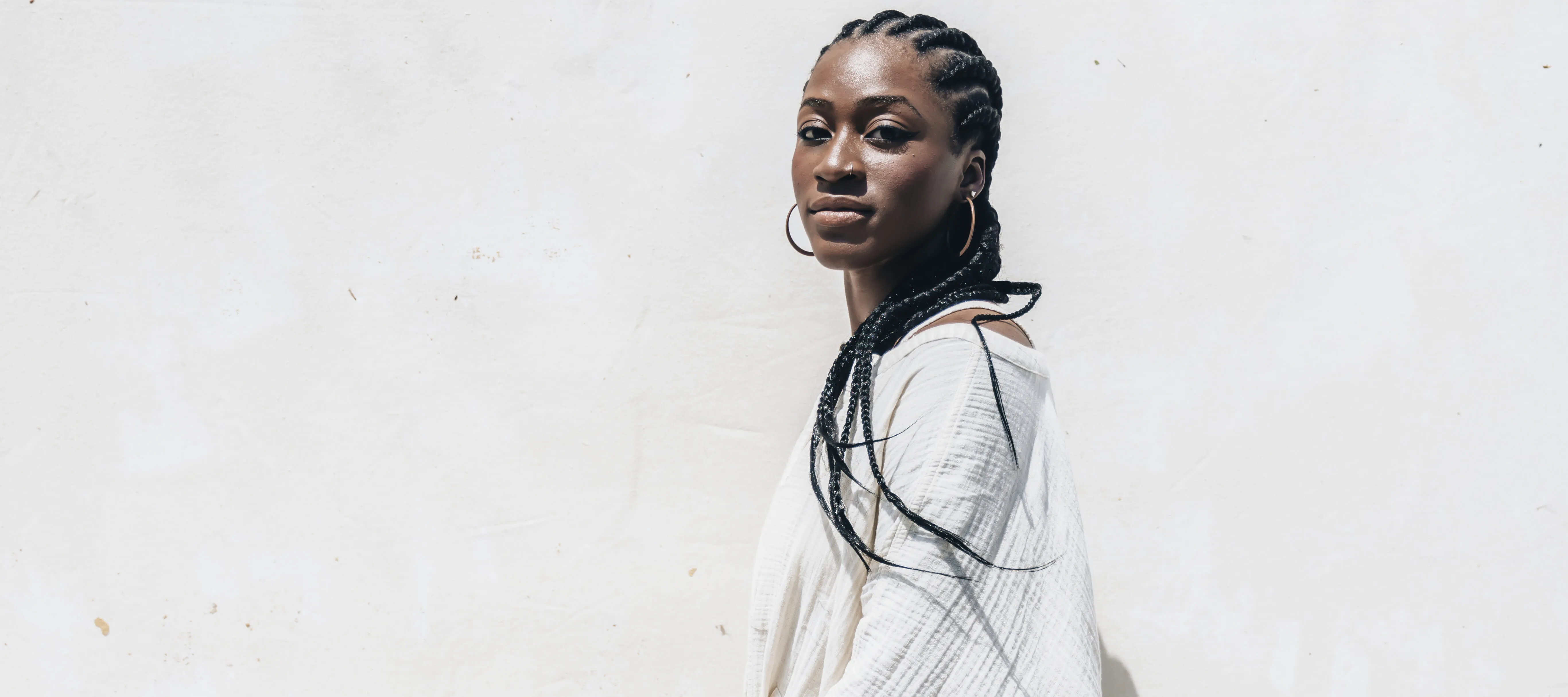 A dark-skinned woman wearing all white sits on a white box against a white background. She looks at the camera confidently.