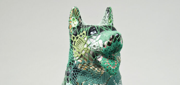 A mass-produced, ceramic German shepherd sits upright and alert with mouth open and tongue extended as if panting. Panels of elaborate hand-crafted crochet in shades of mint, citron, and emerald green form a skin-tight web that entirely envelops the dog.