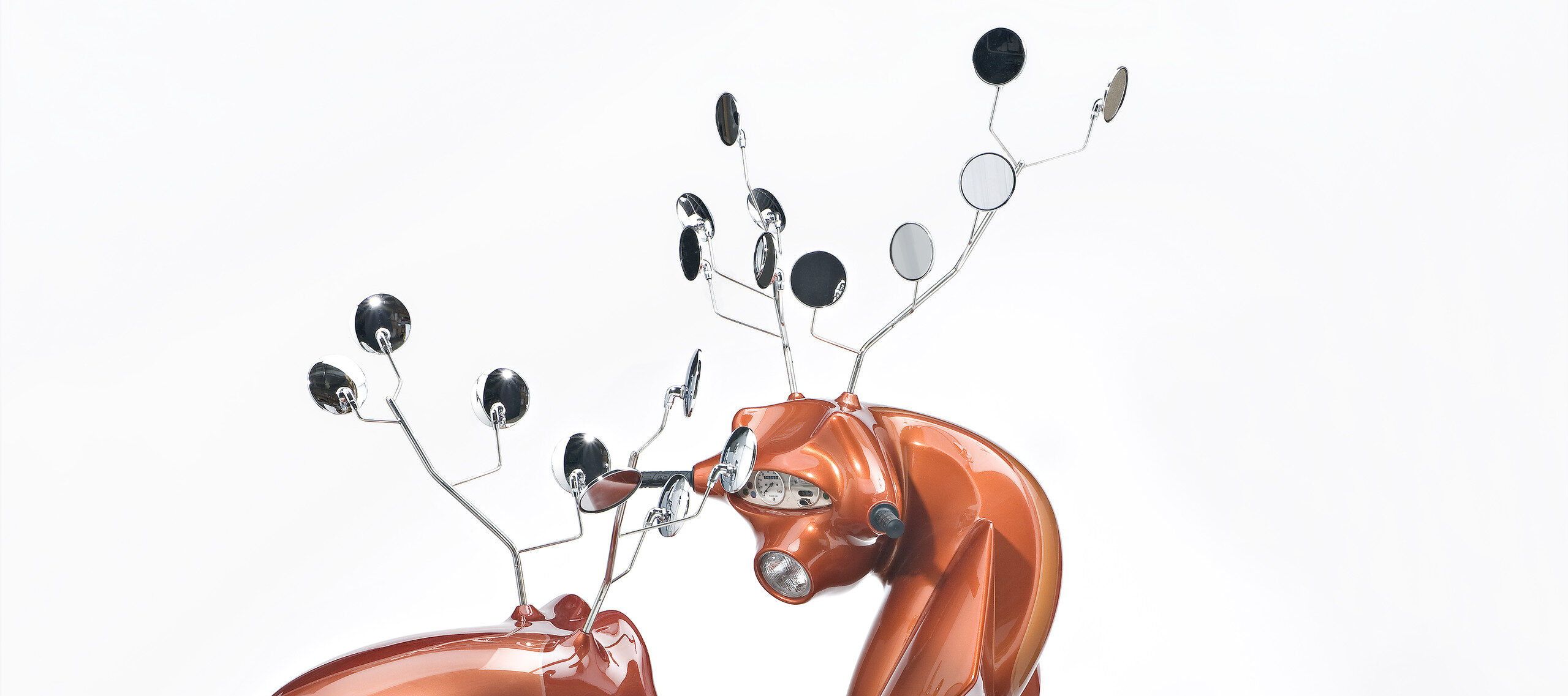 A sculpture consists of two metallic-orange motor scooters manipulated to resemble male deer. Leather seats become haunches, dashboard dials resemble faces, and multiple rear-view mirrors morph into antlers. The serpentine, hybrid animal-machines appear to spar for dominance.