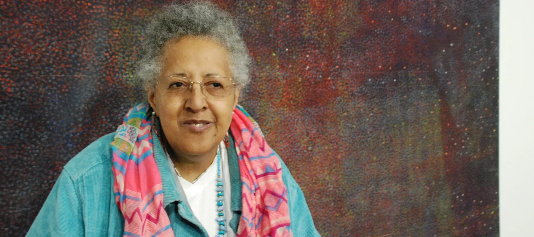 An older woman with medium-dark skin tone smiles slightly while standing in front of an abstract painting. She wears a colorful scarf atop a light-blue corduroy jacket.