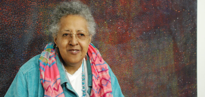 An older woman with medium-dark skin tone smiles slightly while standing in front of an abstract painting. She wears a colorful scarf atop a light-blue corduroy jacket.