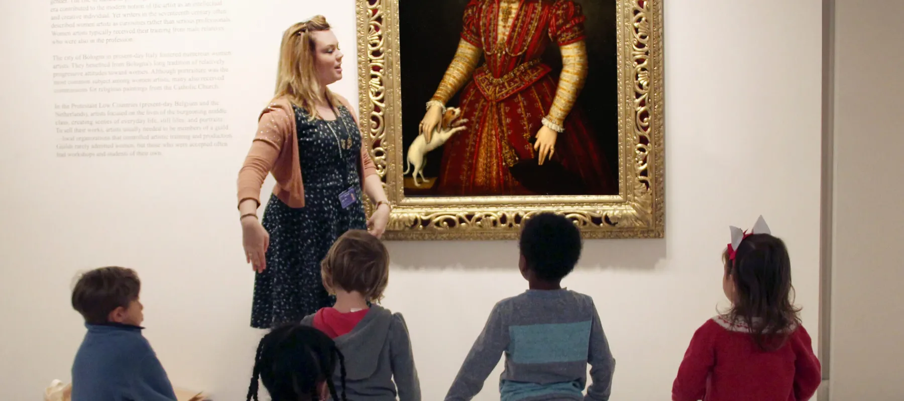 A group of kids standing on colorful mats in a museum space. They are looking towards an oil portrait of a woman in regal garments. A woman with a light skin tone and blonde hair is talking in front of the children.