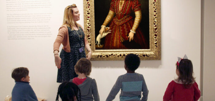 A group of kids standing on colorful mats in a museum space. They are looking towards an oil portrait of a woman in regal garments. A woman with a light skin tone and blonde hair is talking in front of the children.