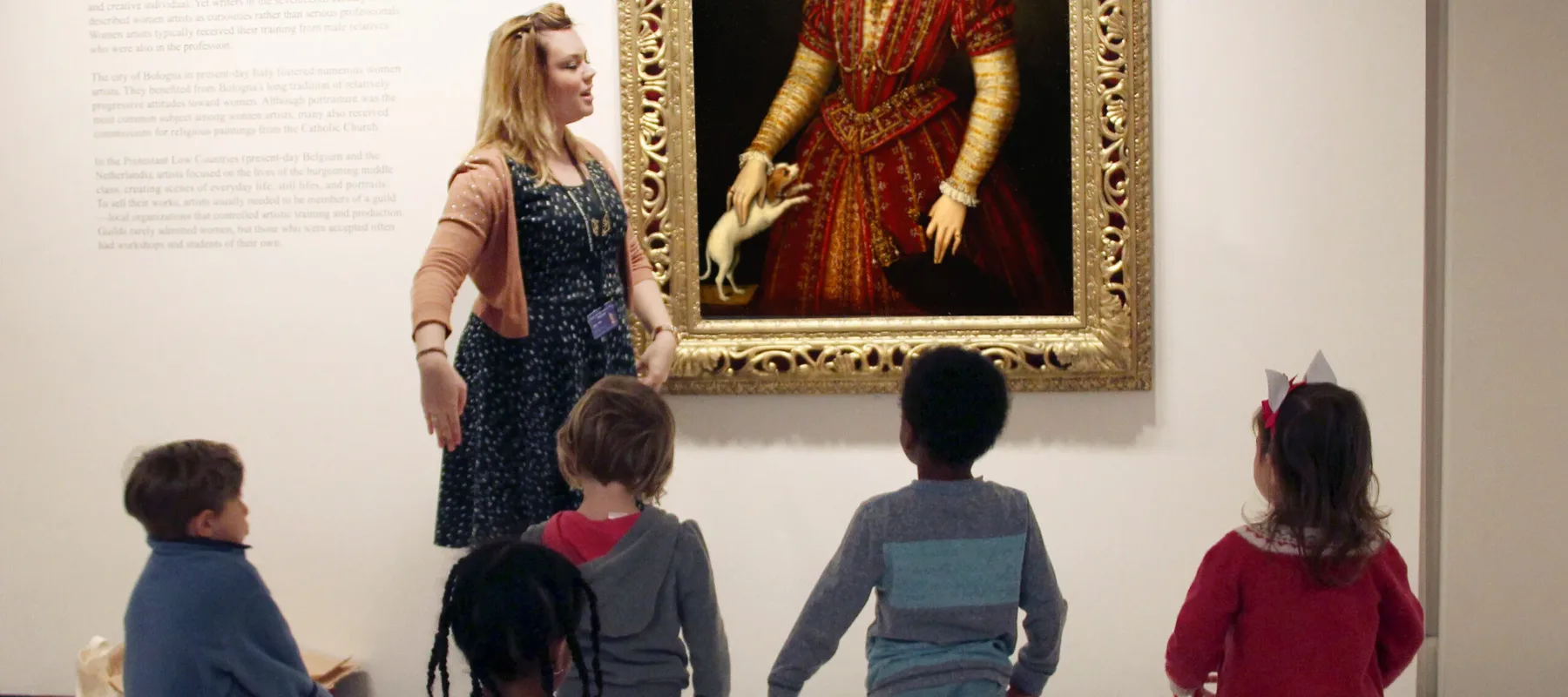 A group of kids standing on colorful mats in a museum space. They are looking towards an oil portrait of a woman in regal garments. A woman with a light skin tone and blonde hair is talking in front of the children.