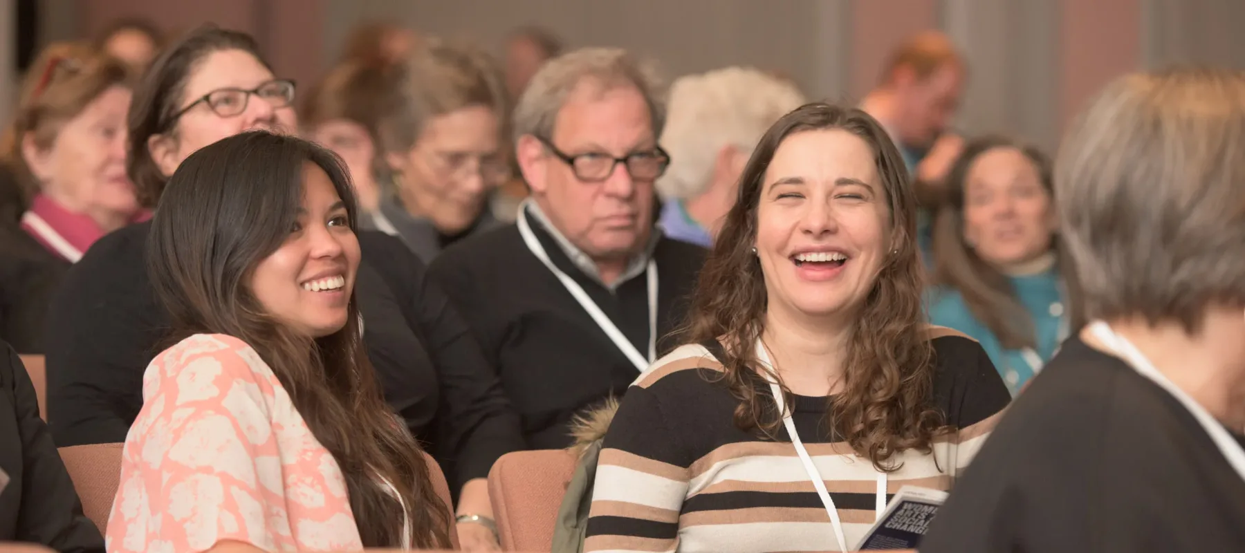 Two light-skinned women with long brown hair sit in a full auditorium. They are both laughing.