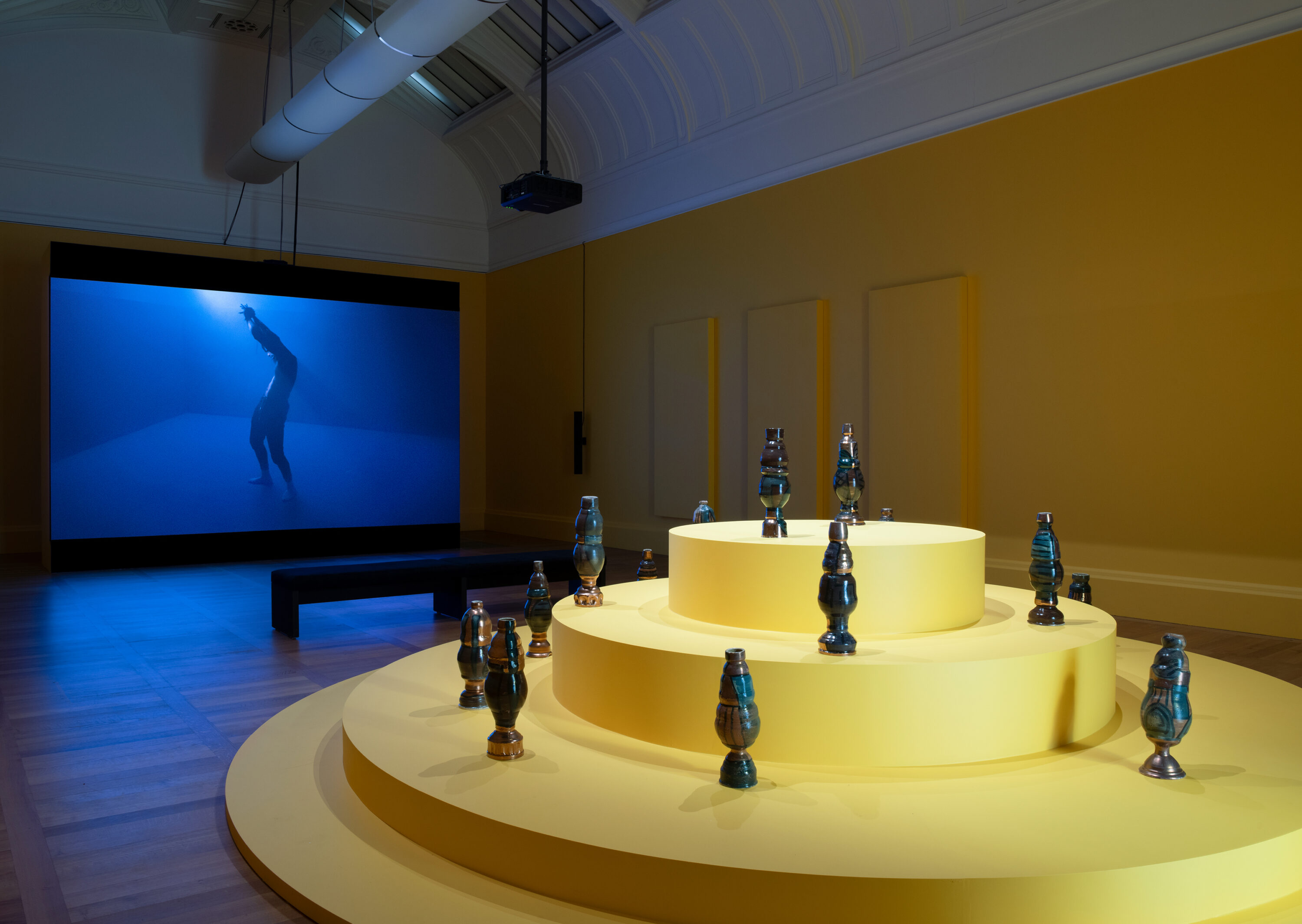 A brightly lit, yellow pedestal composed of four increasingly smaller stacked circles supports multiple thin, vertical ceramic vessels. It occupies the foreground of a barrel-vaulted gallery space with a blue-toned video projected on the back wall. The video shows a single figure in profile posed in the shape of a backward “s.”