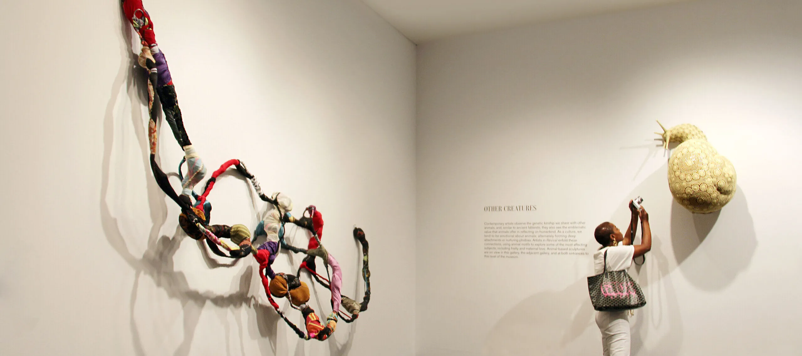 A woman with a dark skin tone wearing an all-white outfit is photographing a sculpture attached to a wall of a snail in a crochet web. Behind her is a large sculpture made from textile, resembling a web-like, string-like object, woven from colorful threads.