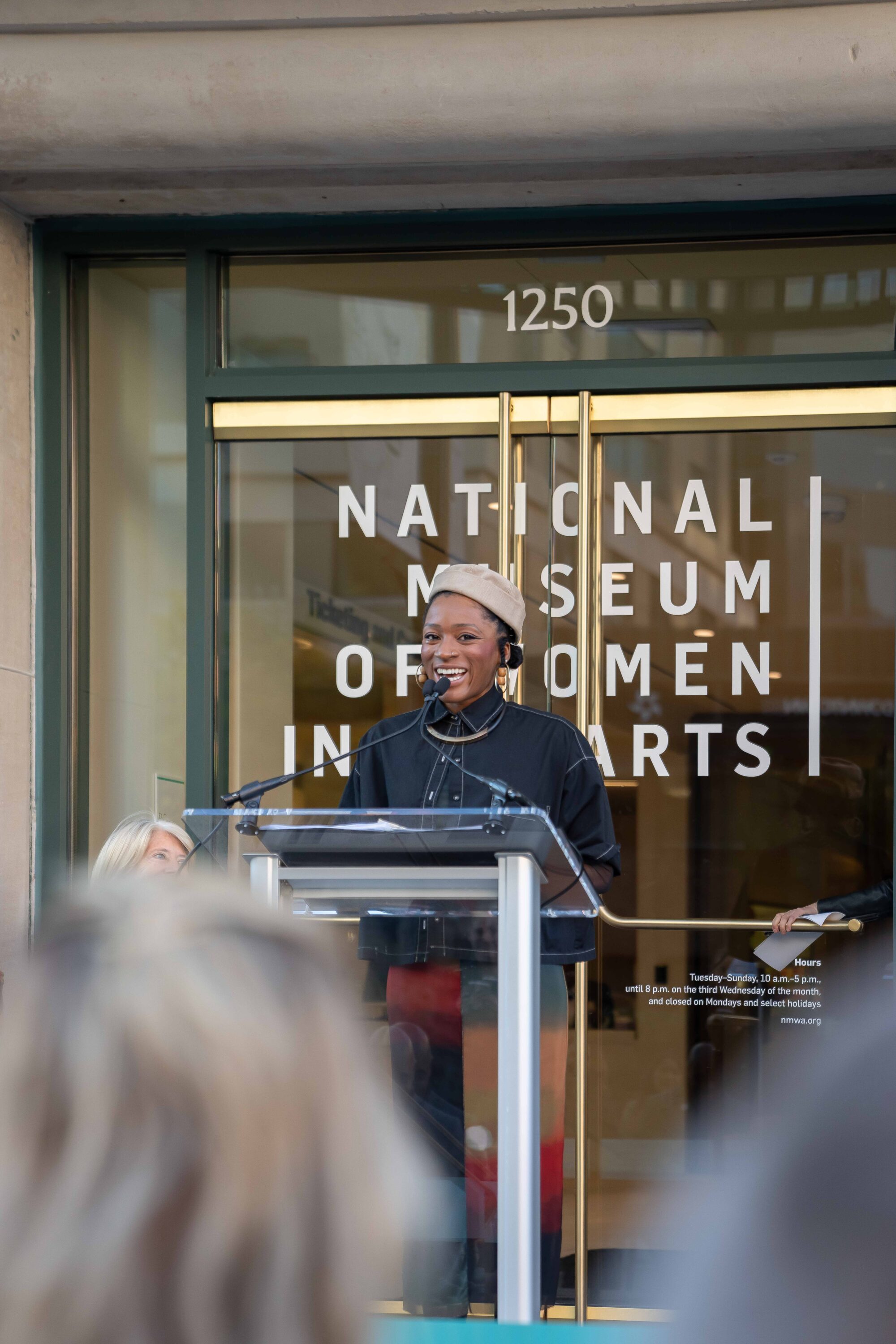 A woman with a dark skin tone wearing a beige beret is giving a speech by a lectern in front of a building. Behind her, it says "National Museum of Women in the Arts" in large, white letters on a glass door.