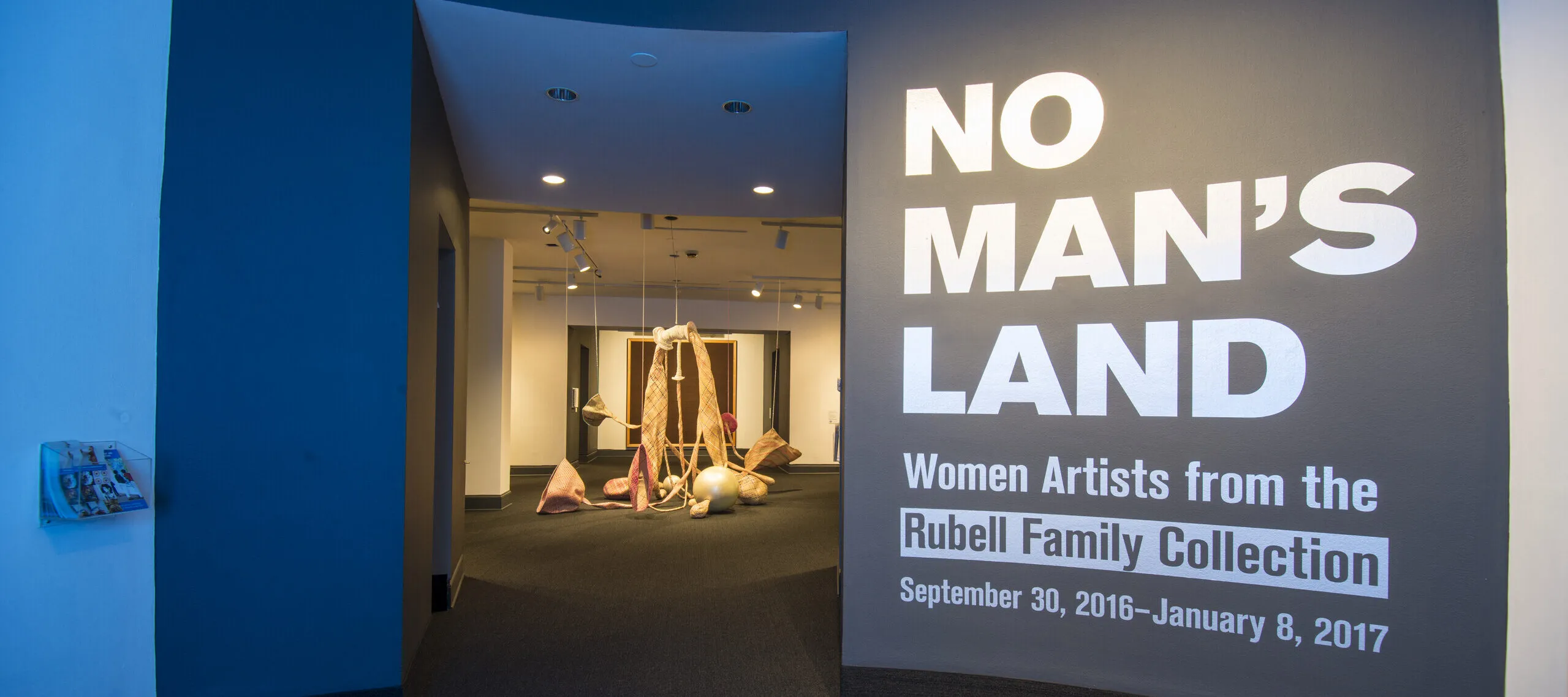 View of a gallery space. On a black wall, it says in white, bold letters: "NO MAN’S LAND: Women Artists from the Rubell Family Collection". There is a textile sculpture hanging from the ceiling in the room behind the wall.