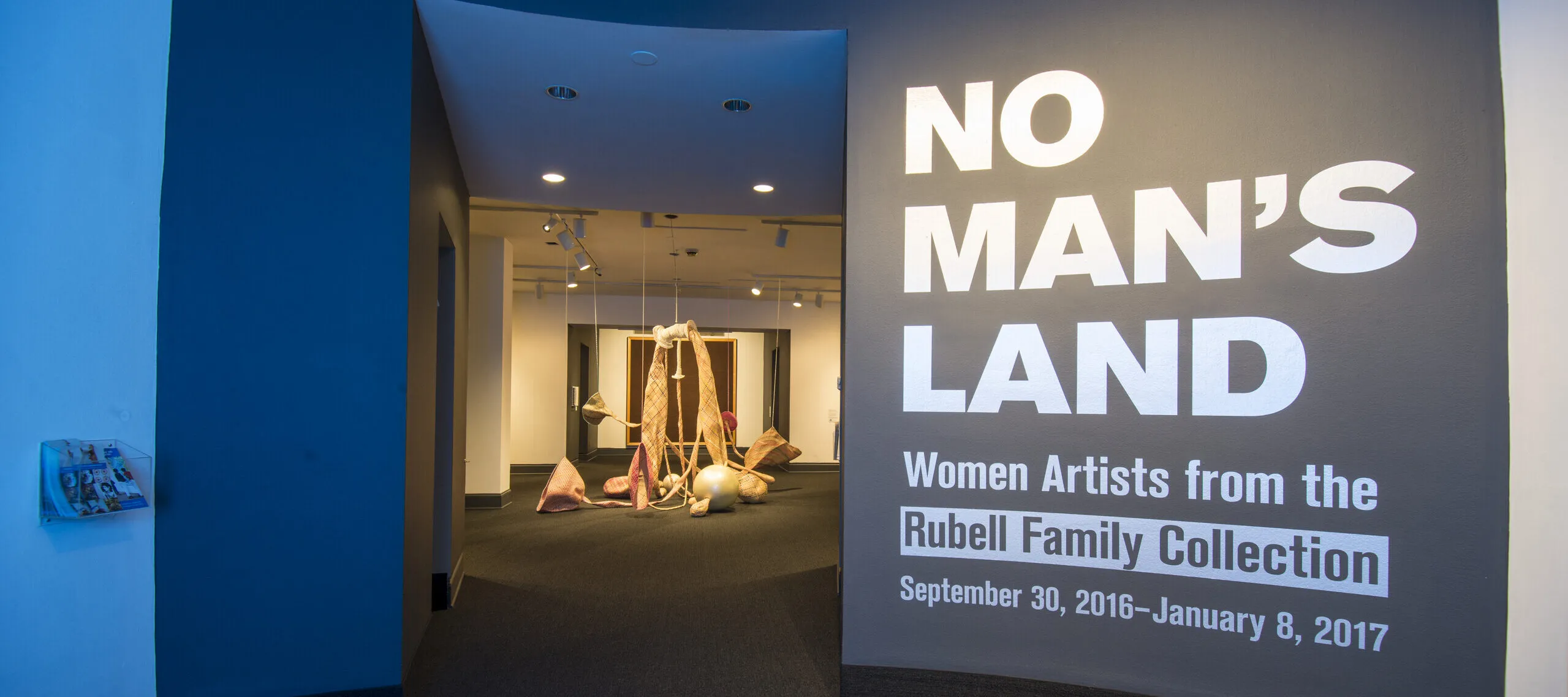 View of a gallery space. On a black wall, it says in white, bold letters: "NO MAN’S LAND: Women Artists from the Rubell Family Collection". There is a textile sculpture hanging from the ceiling in the room behind the wall.