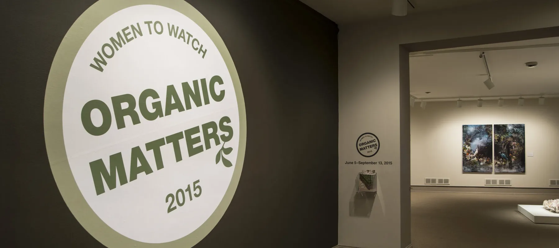 View of a gallery space. In big letters green , the text on the wall reads "Women to Watch: Organic Matters, 2015."