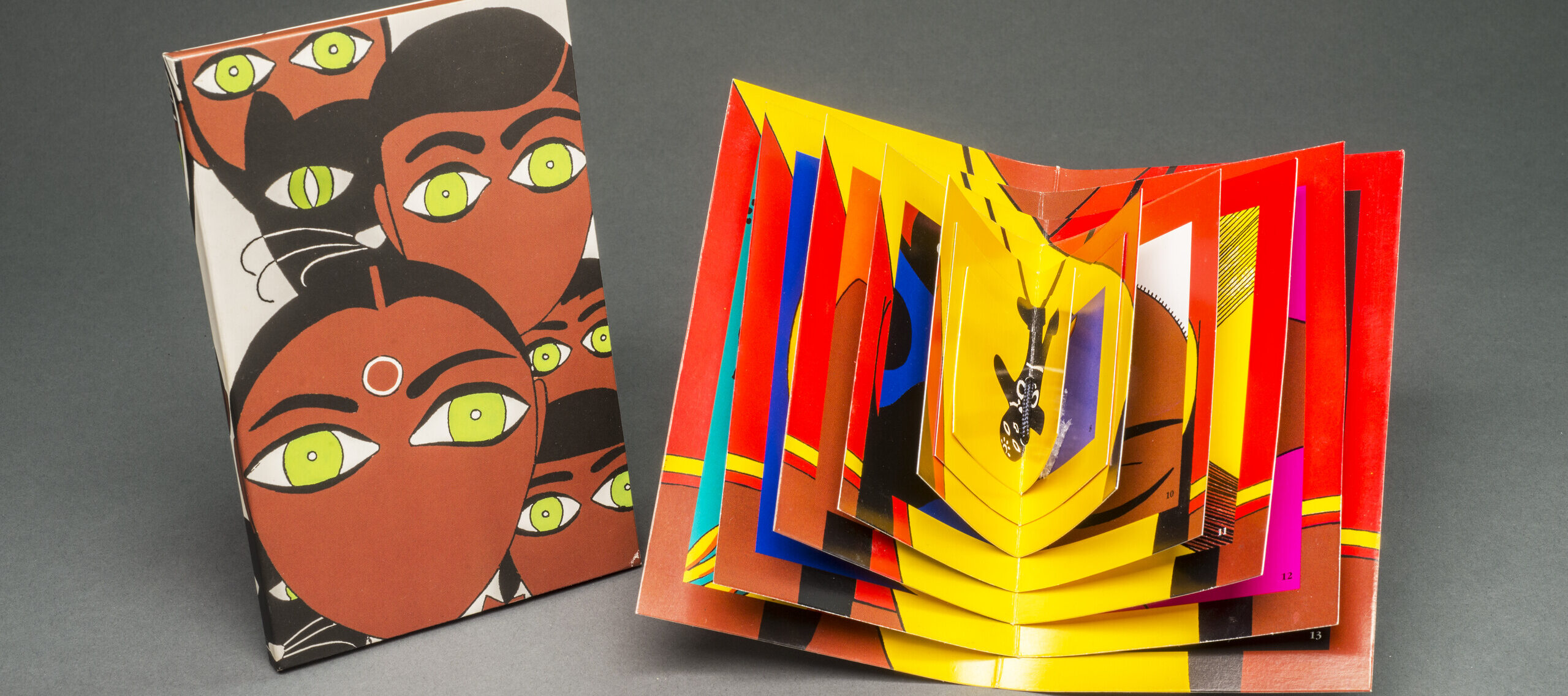 A brightly colored book displayed open whose pages decrease in size toward the center. To the left, a vertical rectangle on which is depicted five stylized faces with black hair, medium-dark skintone,  green eyes, and no mouths. A black cat with green eyes is also among the faces.