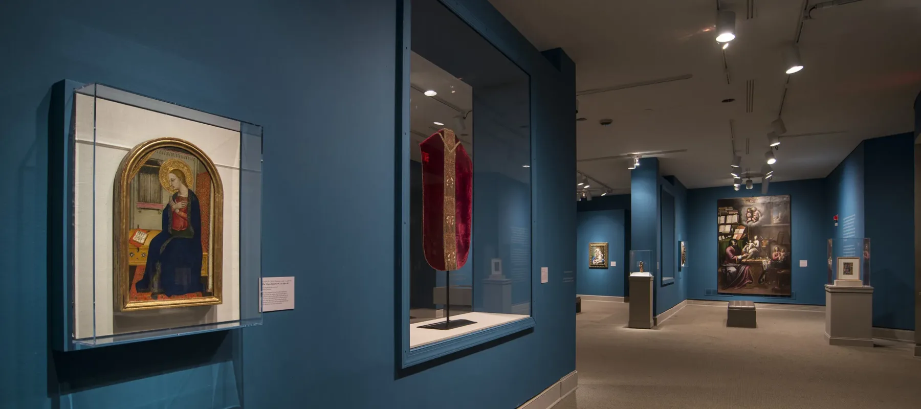 View of a gallery space with blue walls. On the left is a painting of a woman in a dark blue dress in a golden frame. Next to it is a red cloth framed in glass.