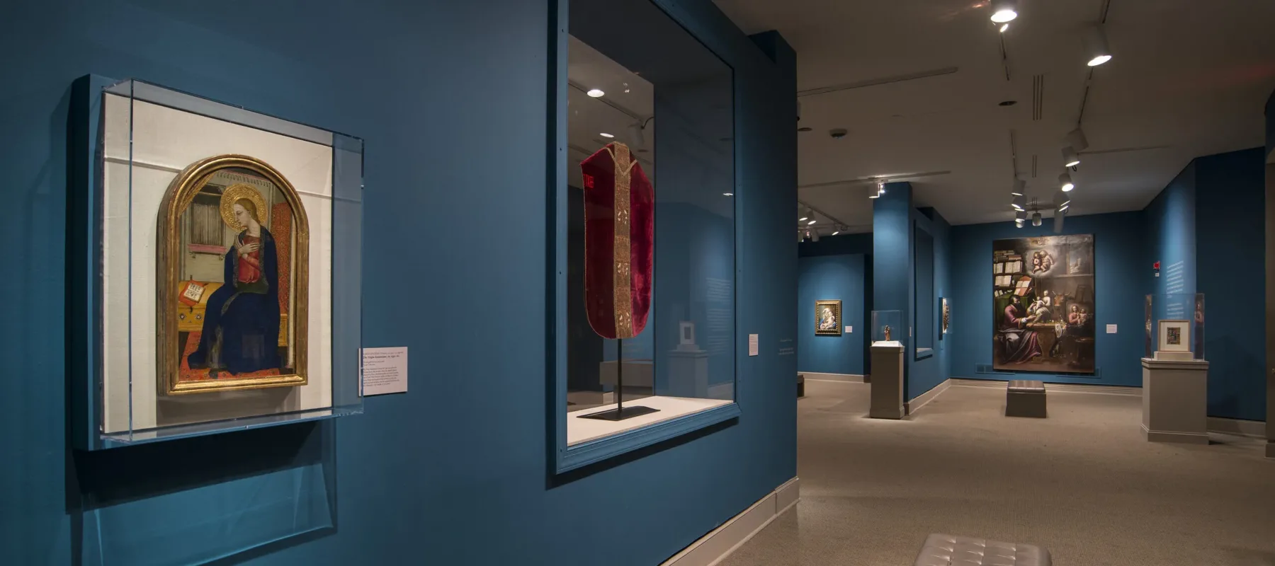 View of a gallery space with blue walls. On the left is a painting of a woman in a dark blue dress in a golden frame. Next to it is a red cloth framed in glass.