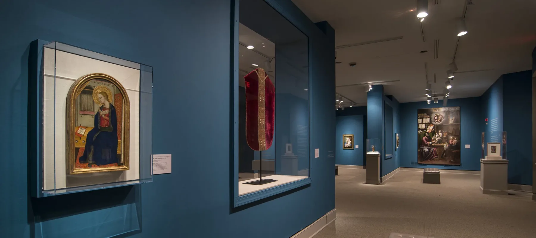 View of a gallery space with blue walls. On the left is a painting of a woman in a dark blue dress in a golden frame. Next to it is a red cloth framed in glass.