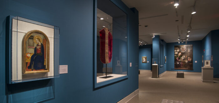 View of a gallery space with blue walls. On the left is a painting of a woman in a dark blue dress in a golden frame. Next to it is a red cloth framed in glass.