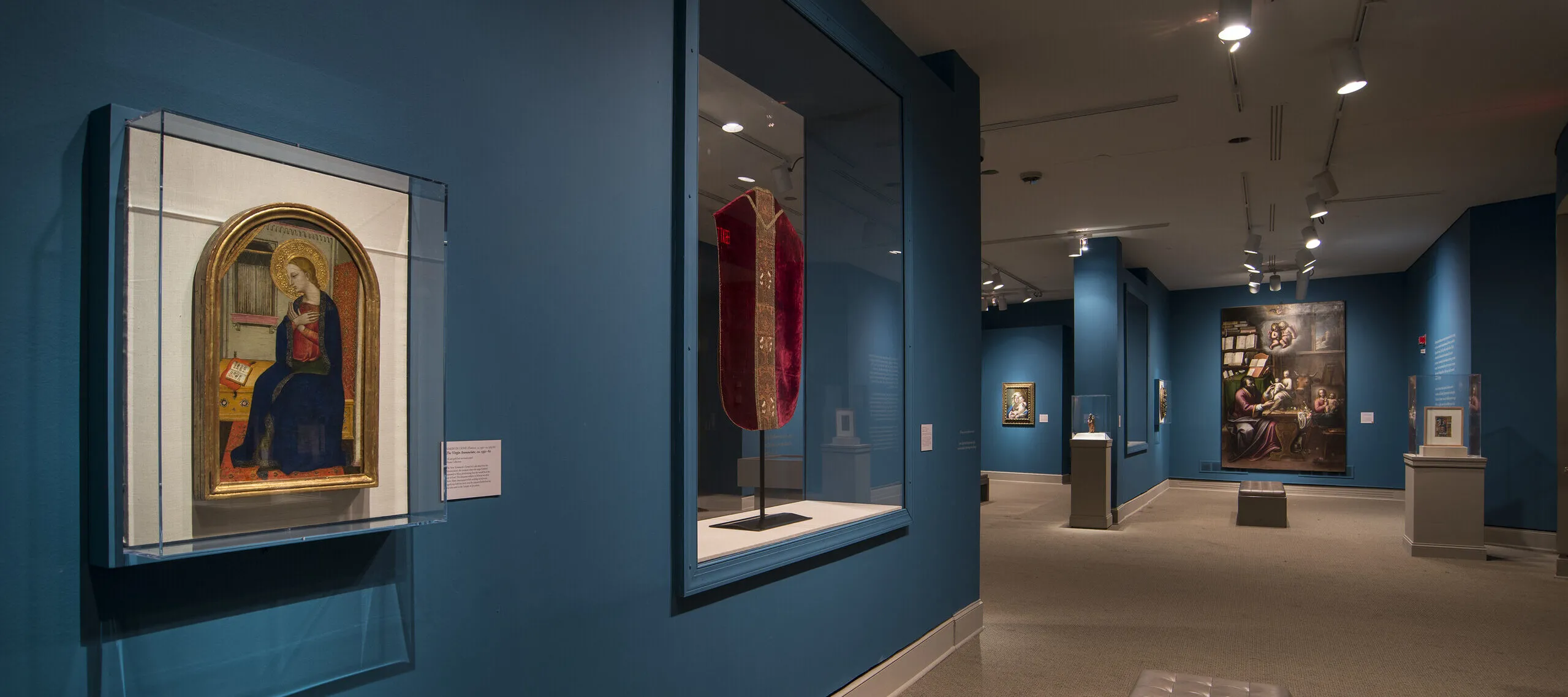 View of a gallery space with blue walls. On the left is a painting of a woman in a dark blue dress in a golden frame. Next to it is a red cloth framed in glass.