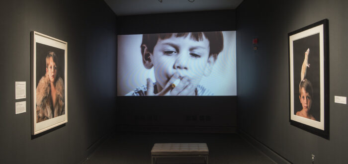 A video screen showing a little boy with a light skin tone smoking is framed by two photographs showing the same boy wearing a fur and with a rat on his head.