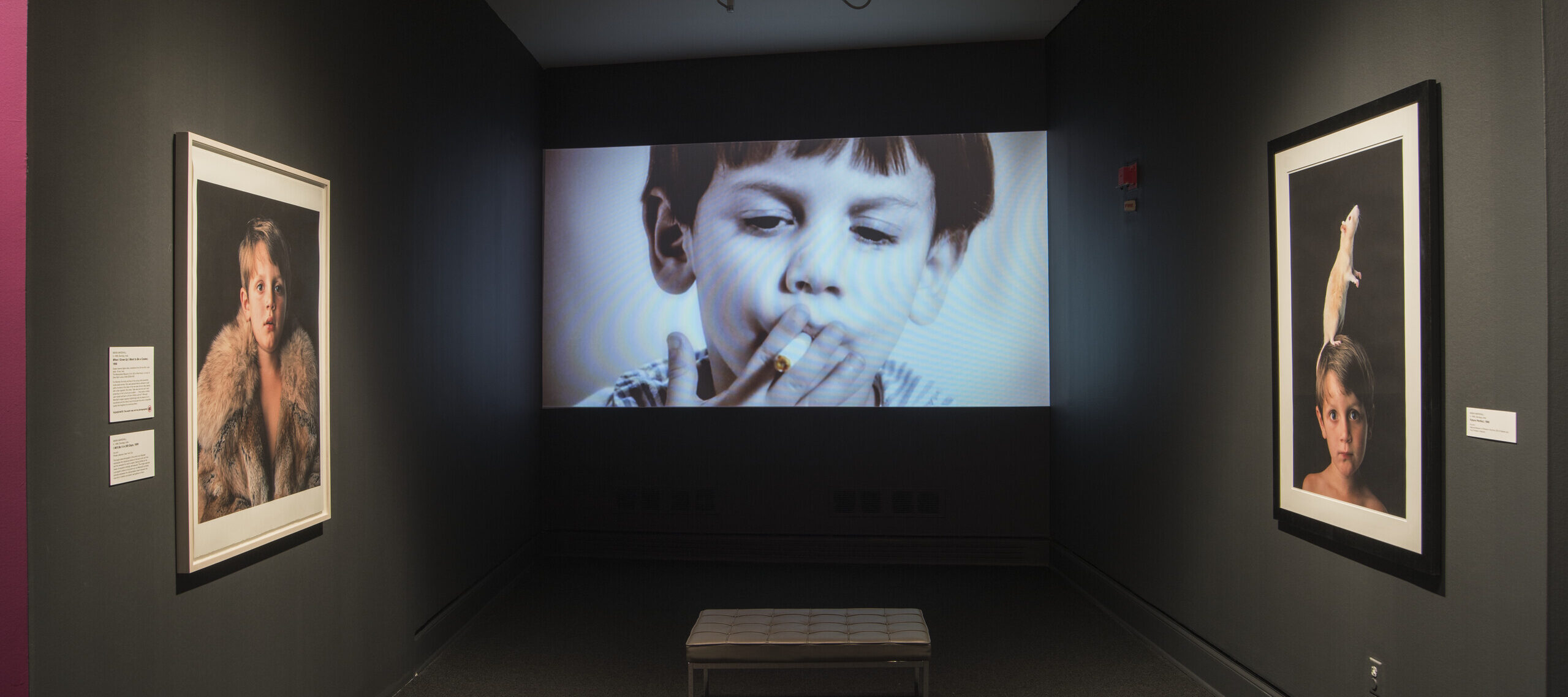 A video screen showing a little boy with a light skin tone smoking is framed by two photographs showing the same boy wearing a fur and with a rat on his head.