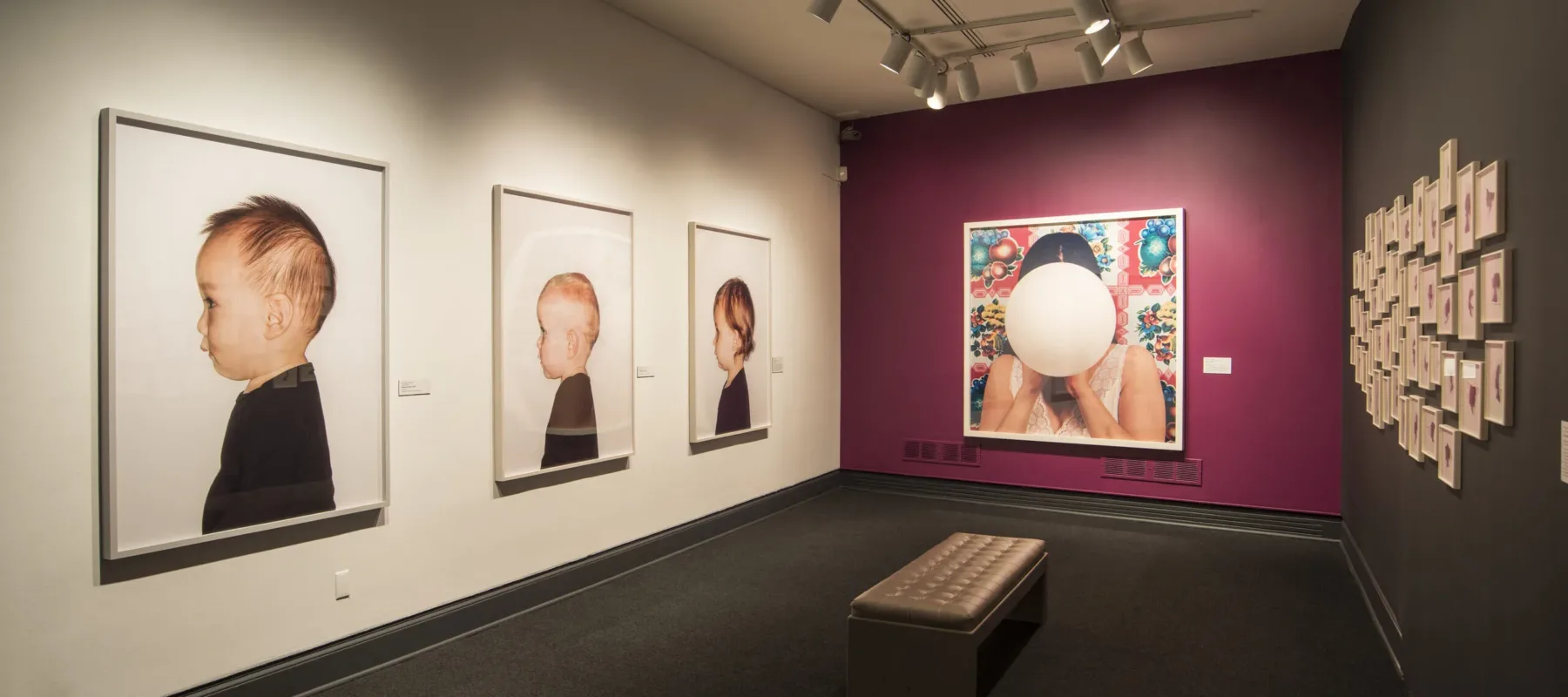 View of a gallery space with a pink and white wall. Three large photographic portraits of babies and young children are hanging next to each other, taking up the whole wall.