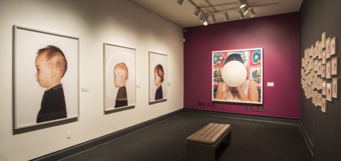 View of a gallery space with a pink and white wall. Three large photographic portraits of babies and young children are hanging next to each other, taking up the whole wall.