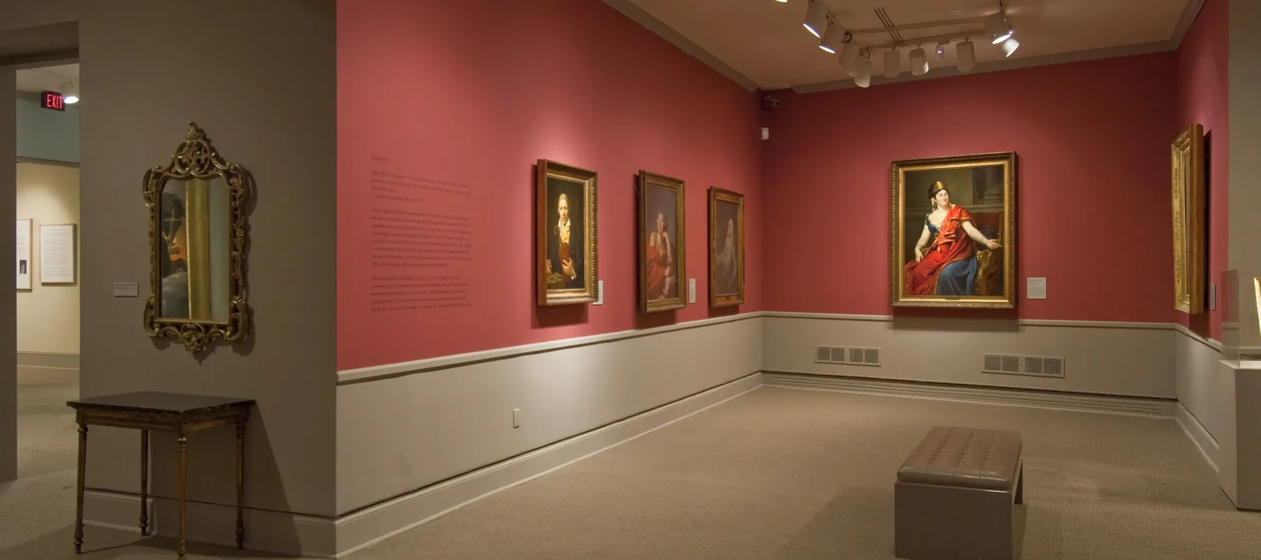 View of a gallery space with red walls. A golden mirror with plenty of ornaments is hanging to the left, large historical portraits of women are hanging to the right.