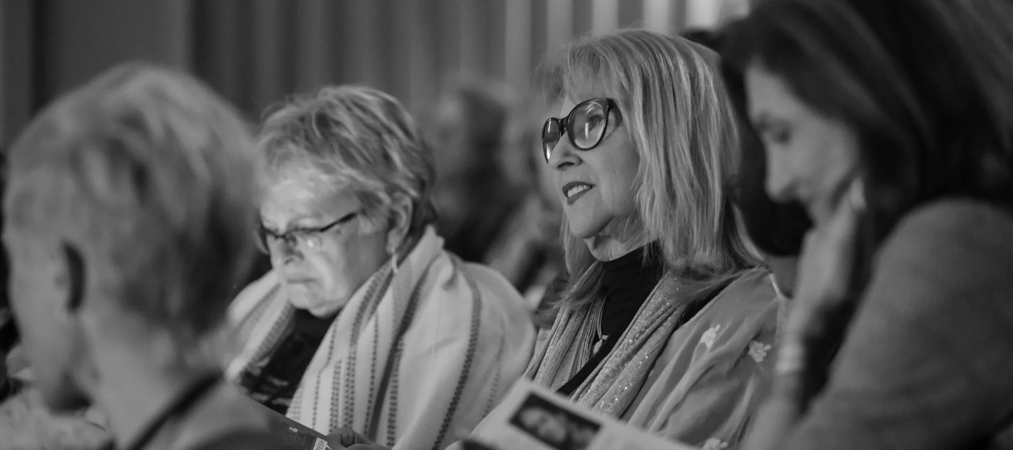 Three light skinned women sit in an auditorium. The woman in the center looks at the stage, the two women on either side of her look down at their programs.