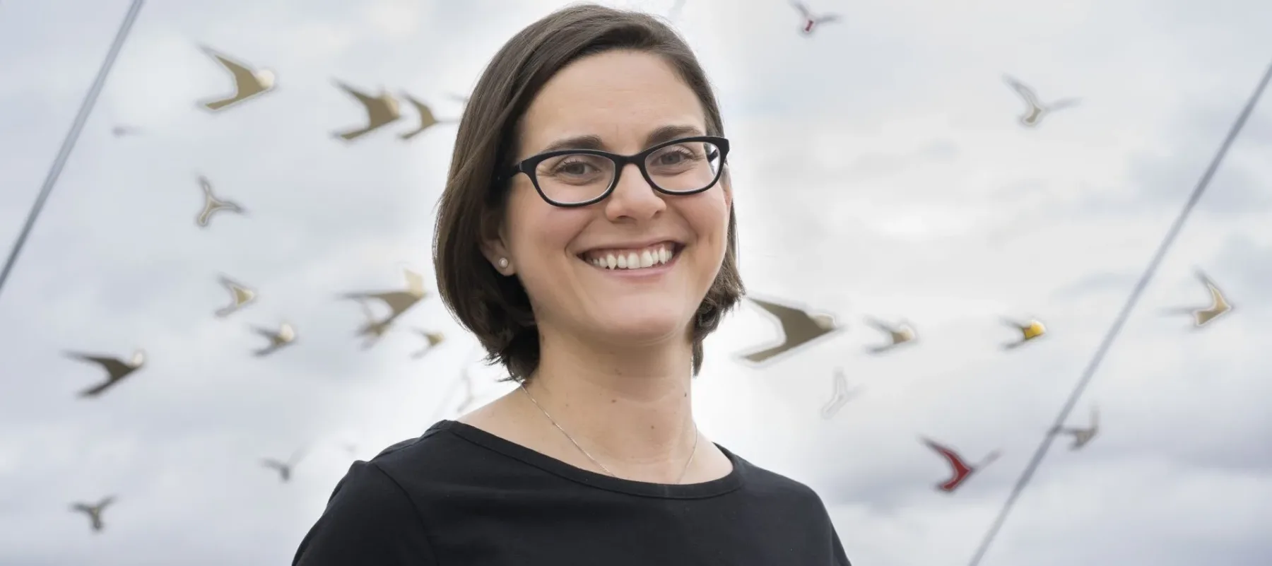 A light-skinned woman is photographed from the chest up. She smiles warmly at the camera against a blue sky with white clouds and what appears to be paper cranes hung on ropes. She wears a black shirt and black-rimmed glasses.