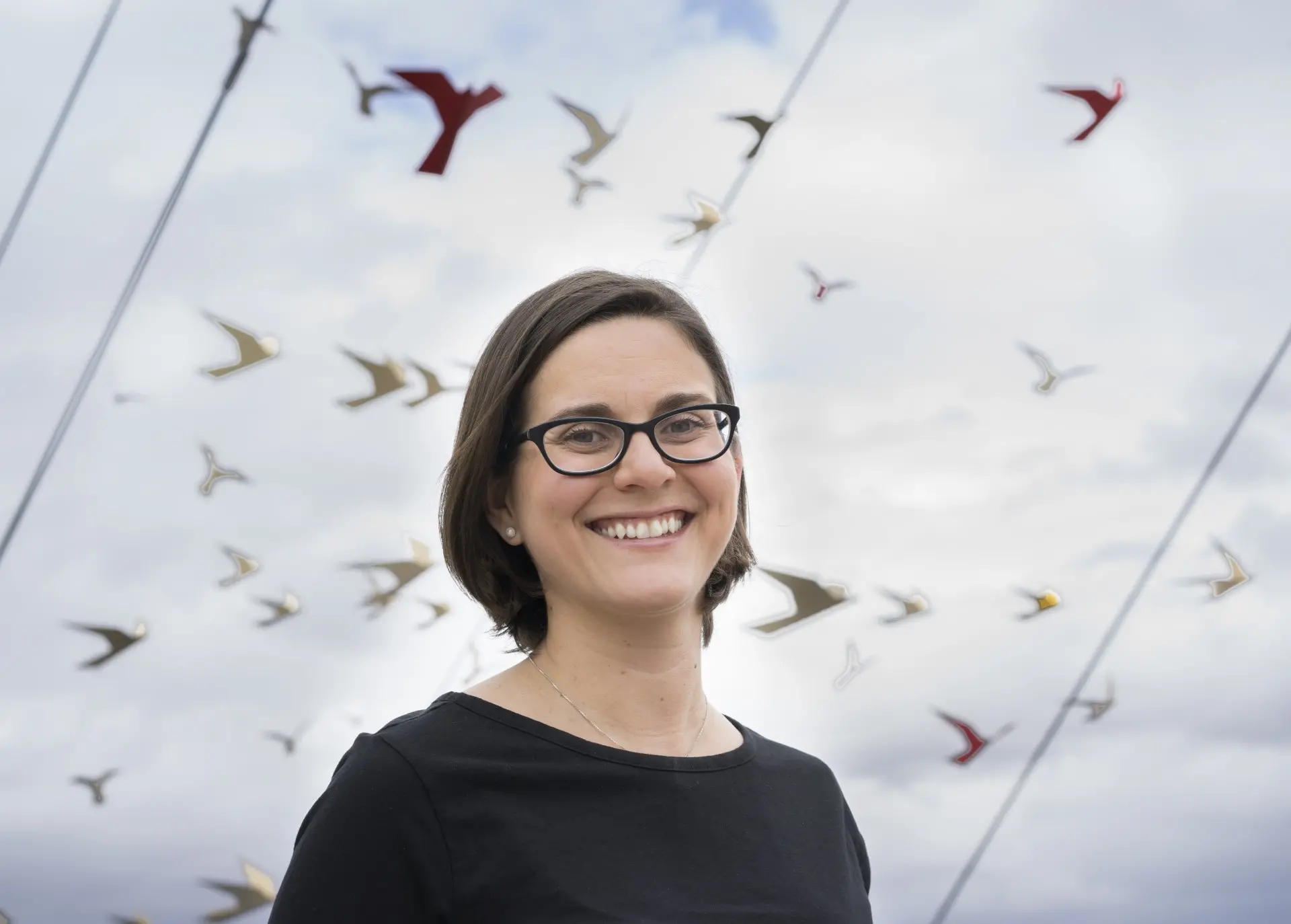 A light-skinned woman is photographed from the chest up. She smiles warmly at the camera against a blue sky with white clouds and what appears to be paper cranes hung on ropes. She wears a black shirt and black-rimmed glasses.