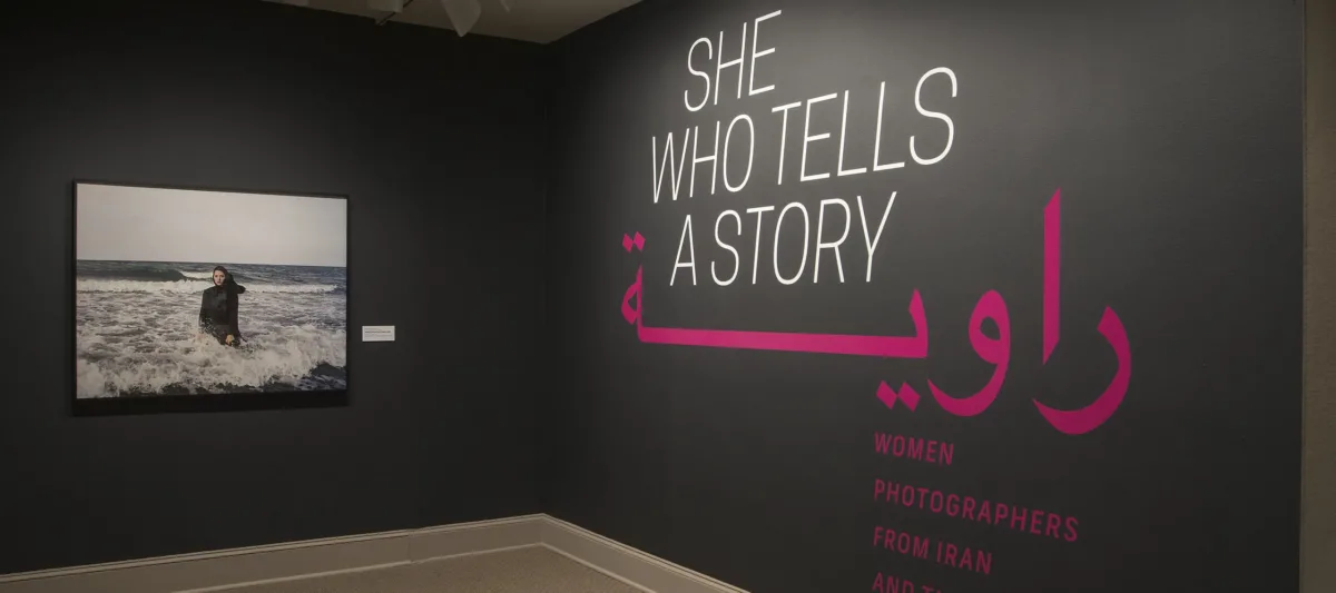 A gallery view of a black wall with a large photograph of a woman. The woman is wearing a long black dress and a head scarf. She is standing in the ocean, surrounded by waves. On the right wall is a text that says "She who tells a story".