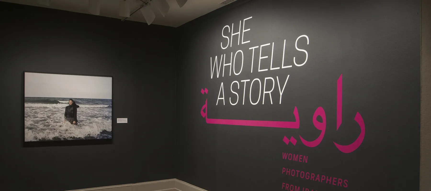 A gallery view of a black wall with a large photograph of a woman. The woman is wearing a long black dress and a head scarf. She is standing in the ocean, surrounded by waves. On the right wall is a text that says "She who tells a story".