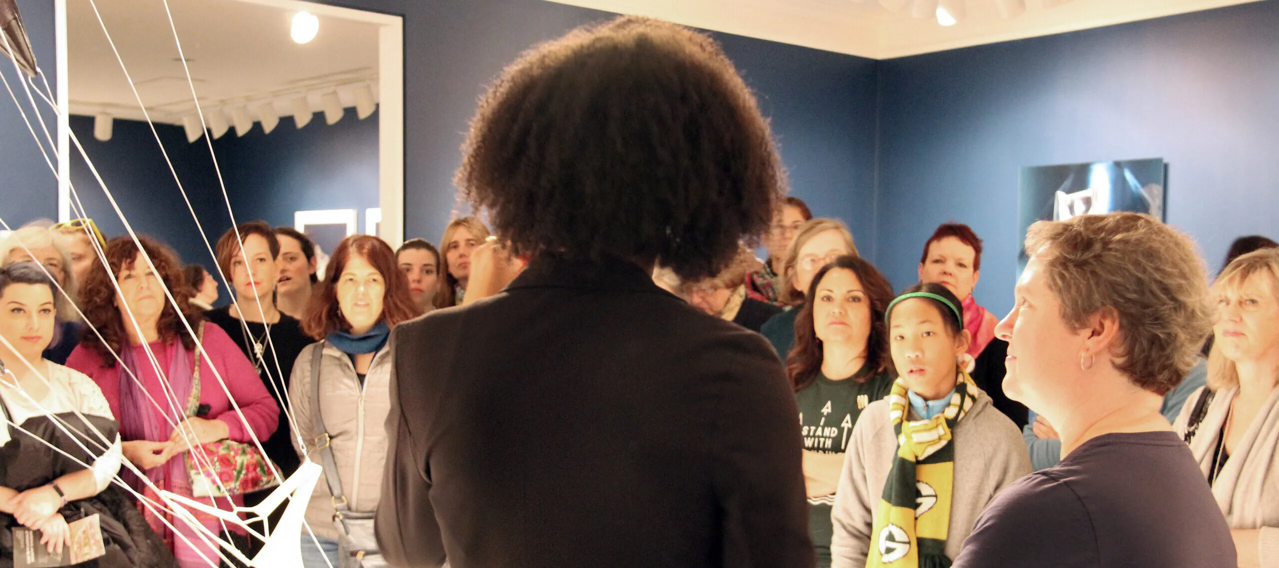 NMWA educator with dark curly hair and a black blazer stands in the foreground with her back to camera as she speaks to a large crowd of visitors about a sculpture composed of hanging strings and fabric in the galleries.